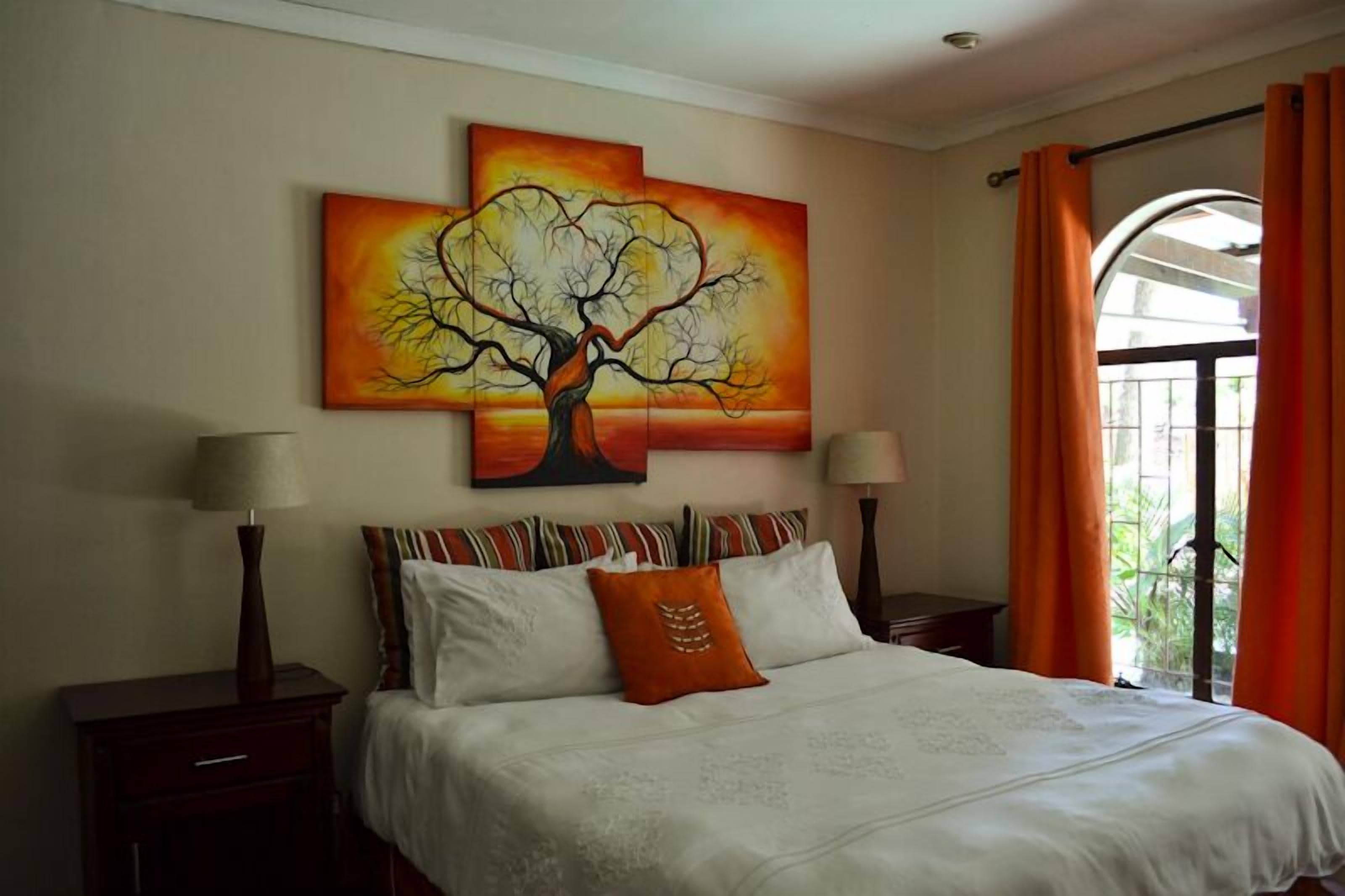 A bedroom with a bed, pillows, a nightstand, a lamp, and a painting of a tree.