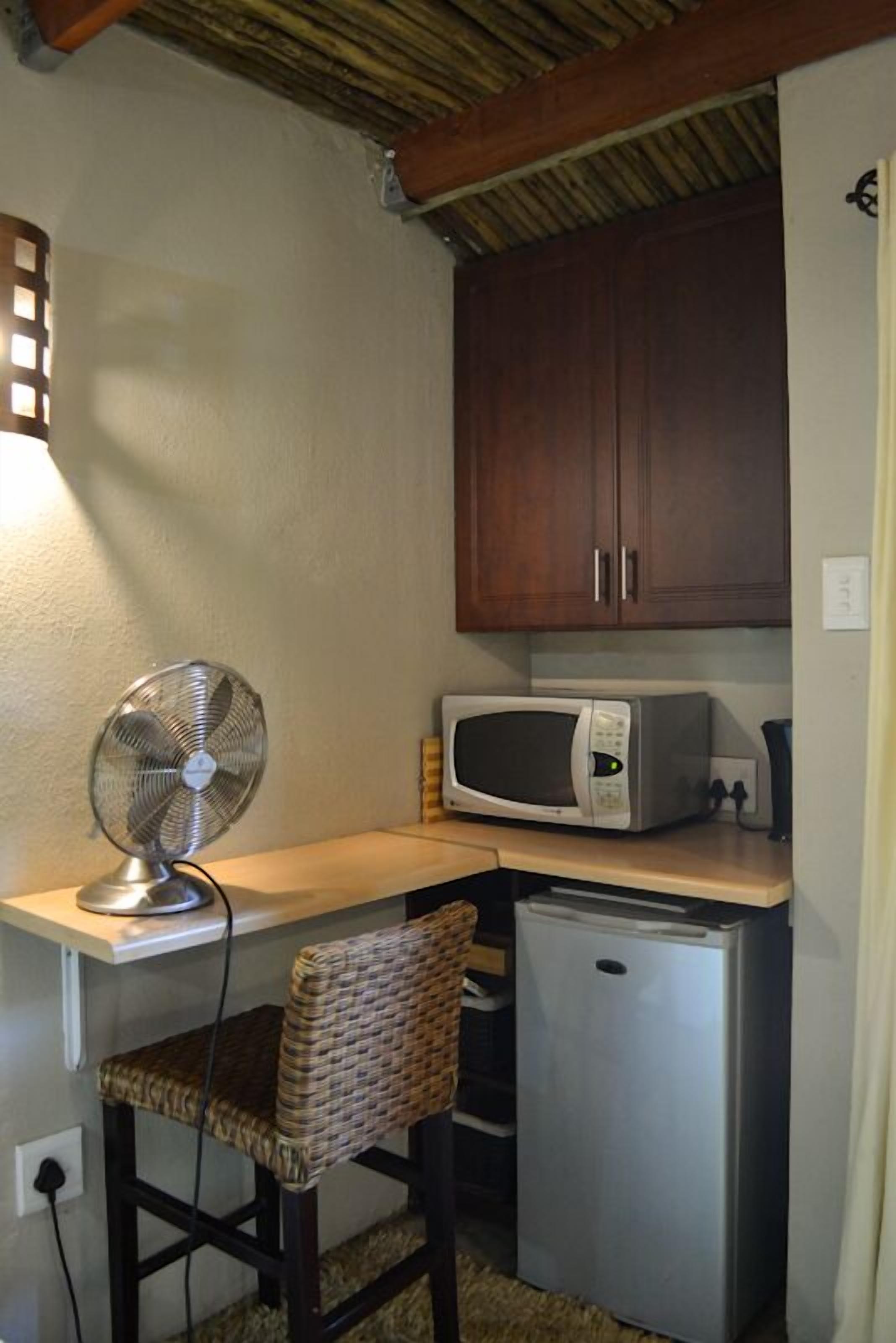 A compact kitchenette with a microwave, a small refrigerator, a fan, and a wicker chair.