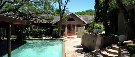 Uma piscina com deck de pedra, um alpendre coberto e uma pequena casa com porta e janelas.