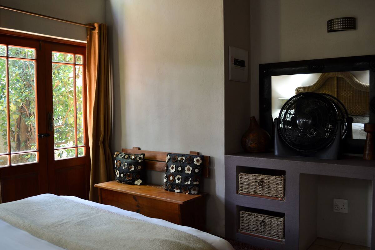 Ein Schlafzimmer mit einem Bett, einer hölzernen Sitzbank mit floralen Kissen, einem großen Ventilator und einem Spiegel an der Wand.