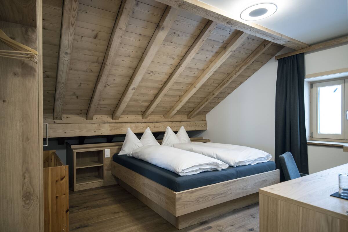 Un dormitorio con techo inclinado, vigas de madera, cama con ropa de cama blanca, un escritorio y una ventana con cortinas.