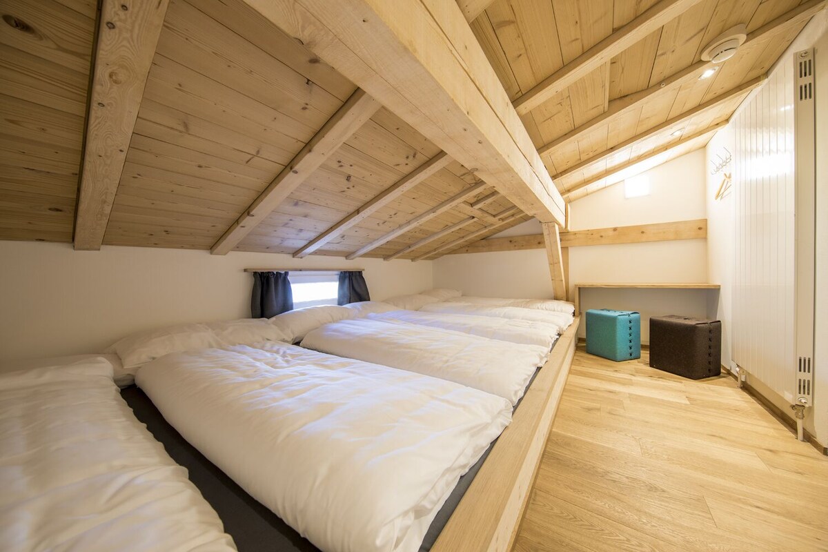 Una habitación con techo inclinado, vigas de madera, una cama con ropa de cama blanca, un ventanal pequeño con cortinas y un radiador.