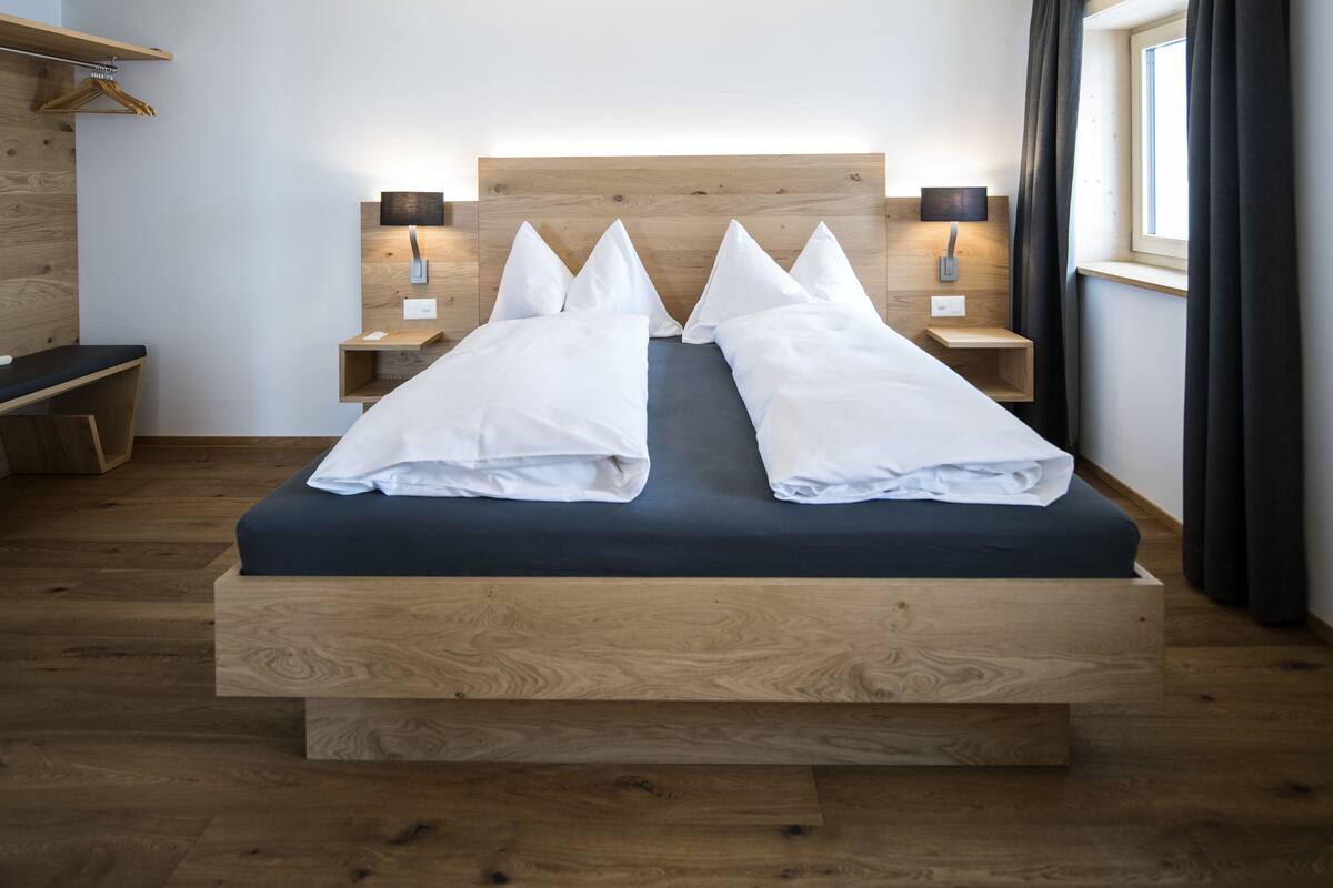 Un dormitorio moderno con cabecera de madera, dos mesitas de noche con lámparas, un banco y una ventana con cortinas.