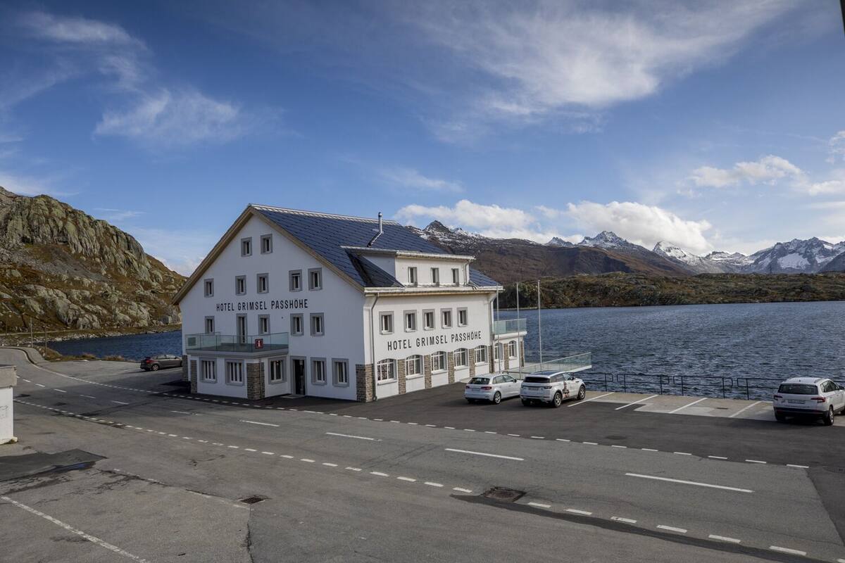 Un edificio blanco con el nombre "Hotel Grimsel Passhöhe" en su fachada, situado junto a un cuerpo de agua con montañas al fondo.