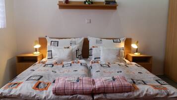 A double bed with a patterned bedspread, two bedside lamps, a wooden shelf with books and flowers, and a bedside table with a lamp.