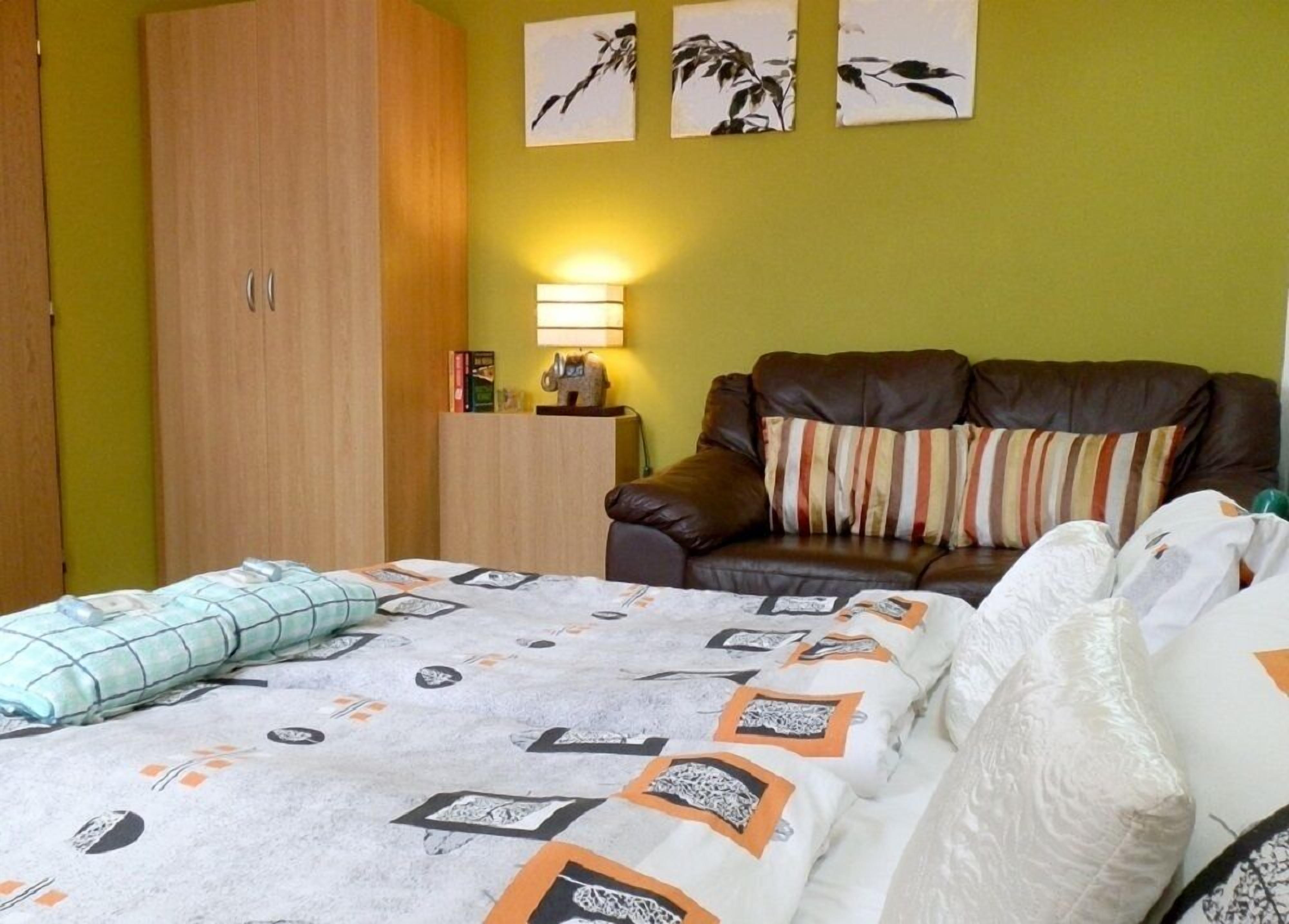 A bedroom with a bed, a brown leather sofa, a wooden wardrobe, and two wall-mounted lamps.