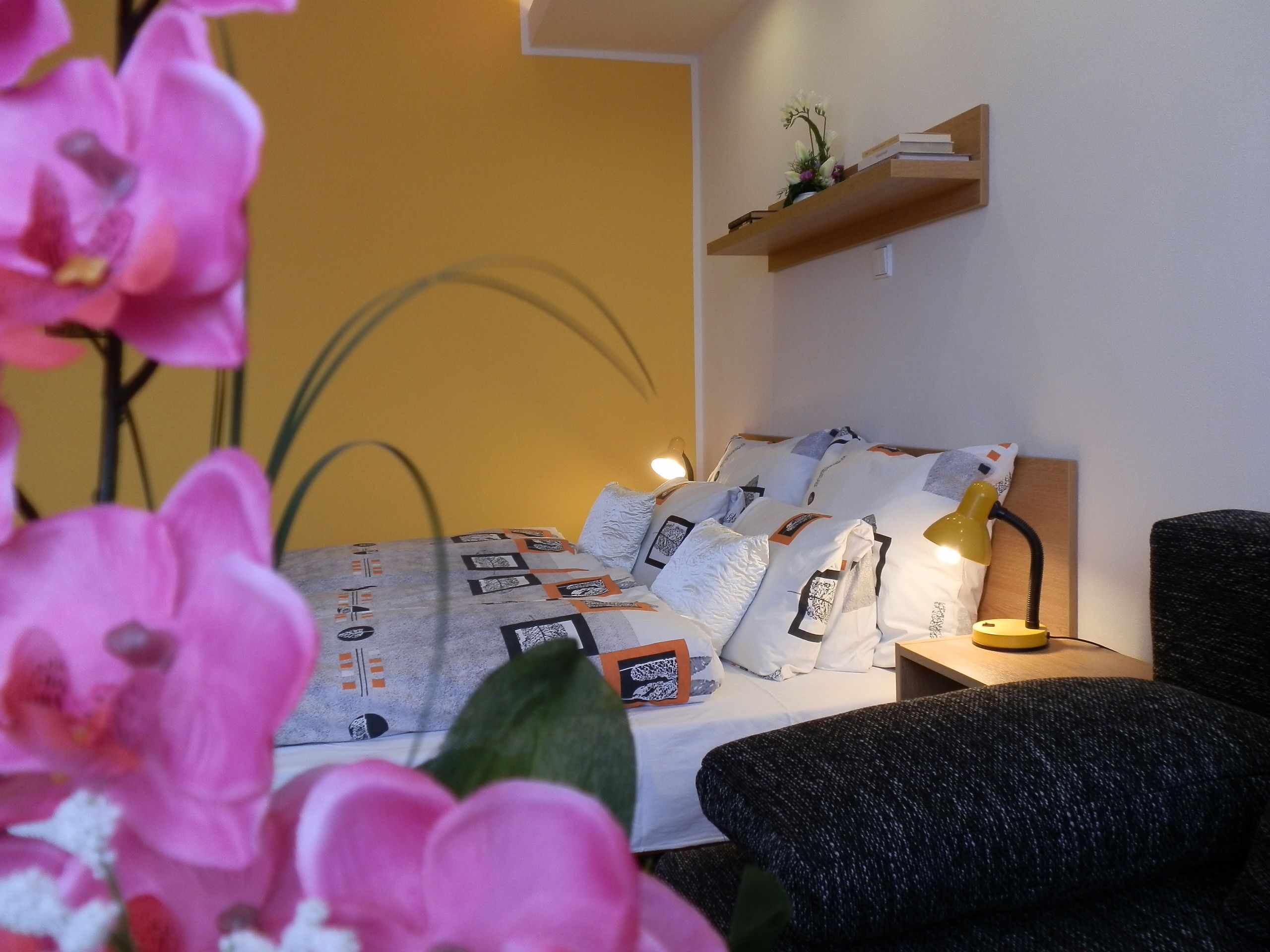 A bedroom with a bed, pillows, a bedside table with a lamp, a shelf with books, and a plant.