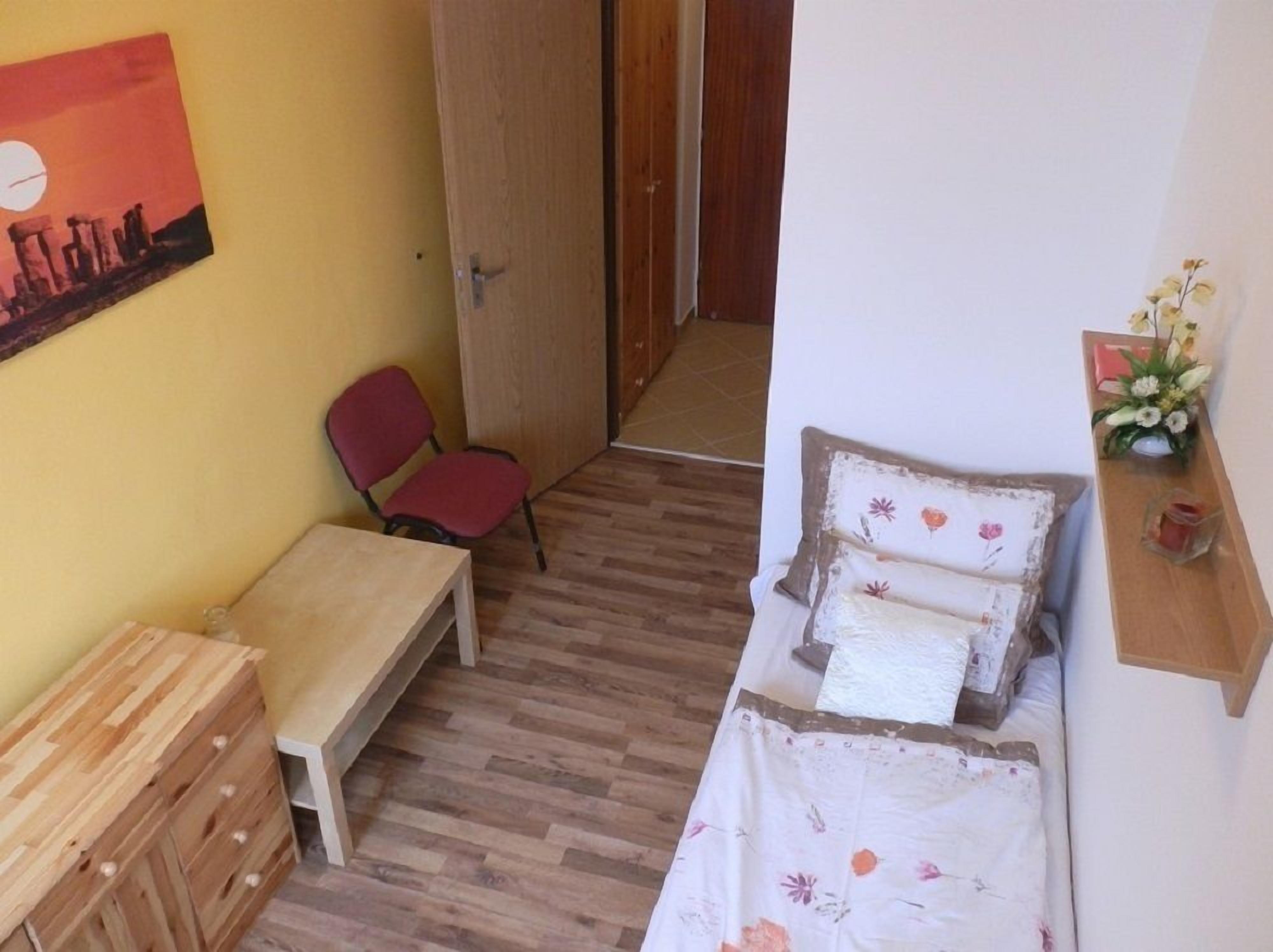 Una habitación pequeña y acogedora con piso de madera, una cama individual con ropa de cama floral, un escritorio pequeño, una silla y un aparador de madera.