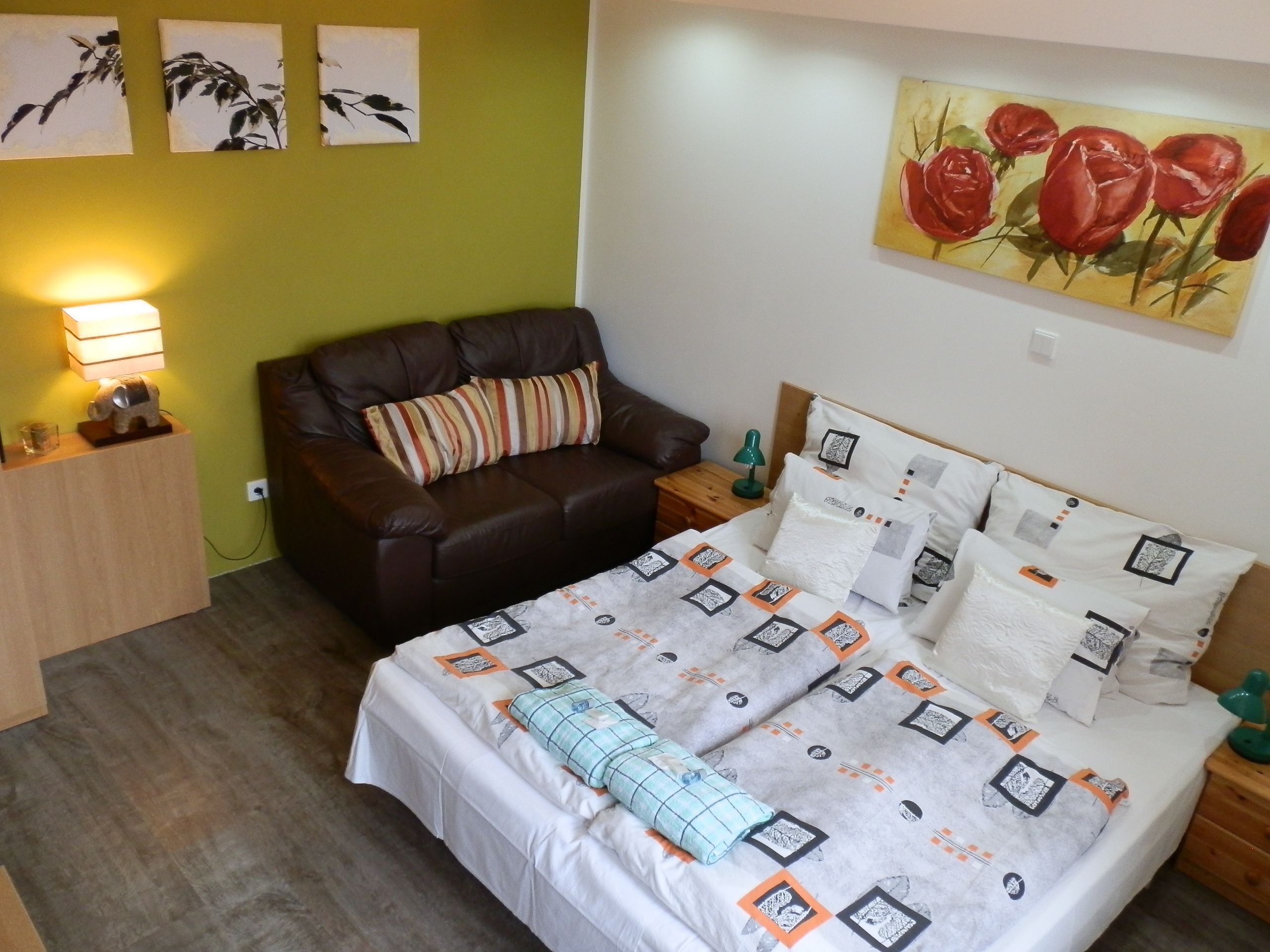 A bedroom with a bed, a brown leather sofa, a wooden nightstand, a green lamp, and a wall with three black and white paintings.
