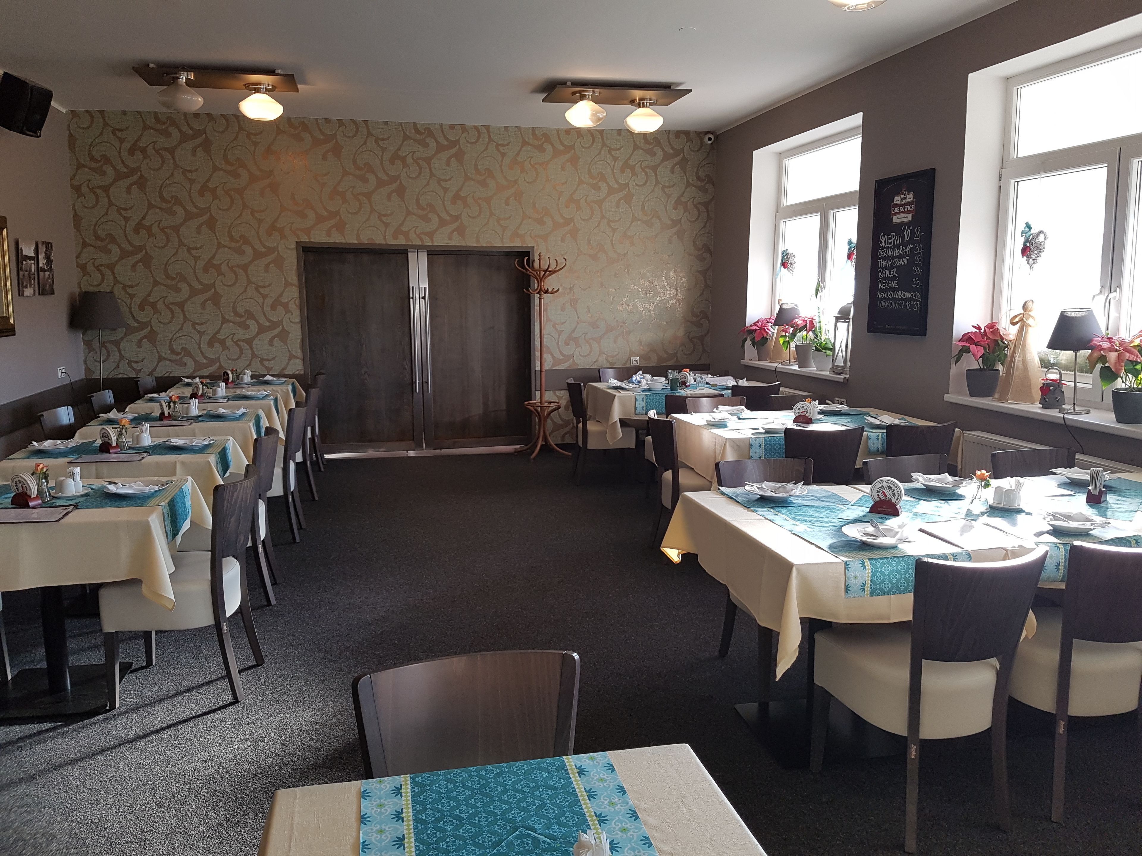 Een restaurant met meerdere gedekte tafels, een grote houten deur en potplanten bij de ramen.