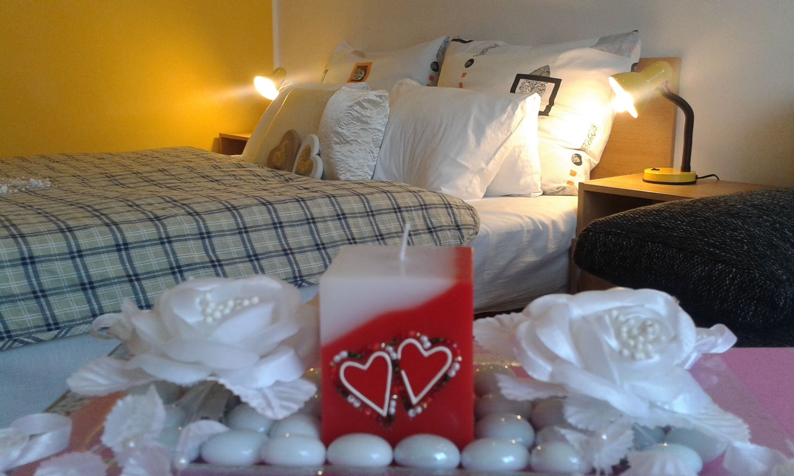 Une chambre d’hôtel avec un lit, des oreillers, une table de chevet avec une lampe, et une composition décorative de fleurs blanches et d’une bougie rouge ornée de motifs en forme de cœur.