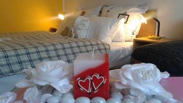 Camera d'albergo con un letto, cuscini, un comodino con una lampada e un'elegante composizione di fiori bianchi e una candela rossa decorata con cuori.