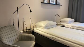 A hotel room with a single bed, a chair, a bedside table with a lamp, and a shelf with framed pictures.