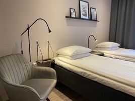 A hotel room with a single bed, a chair, a bedside table with a lamp, and a shelf with framed pictures.