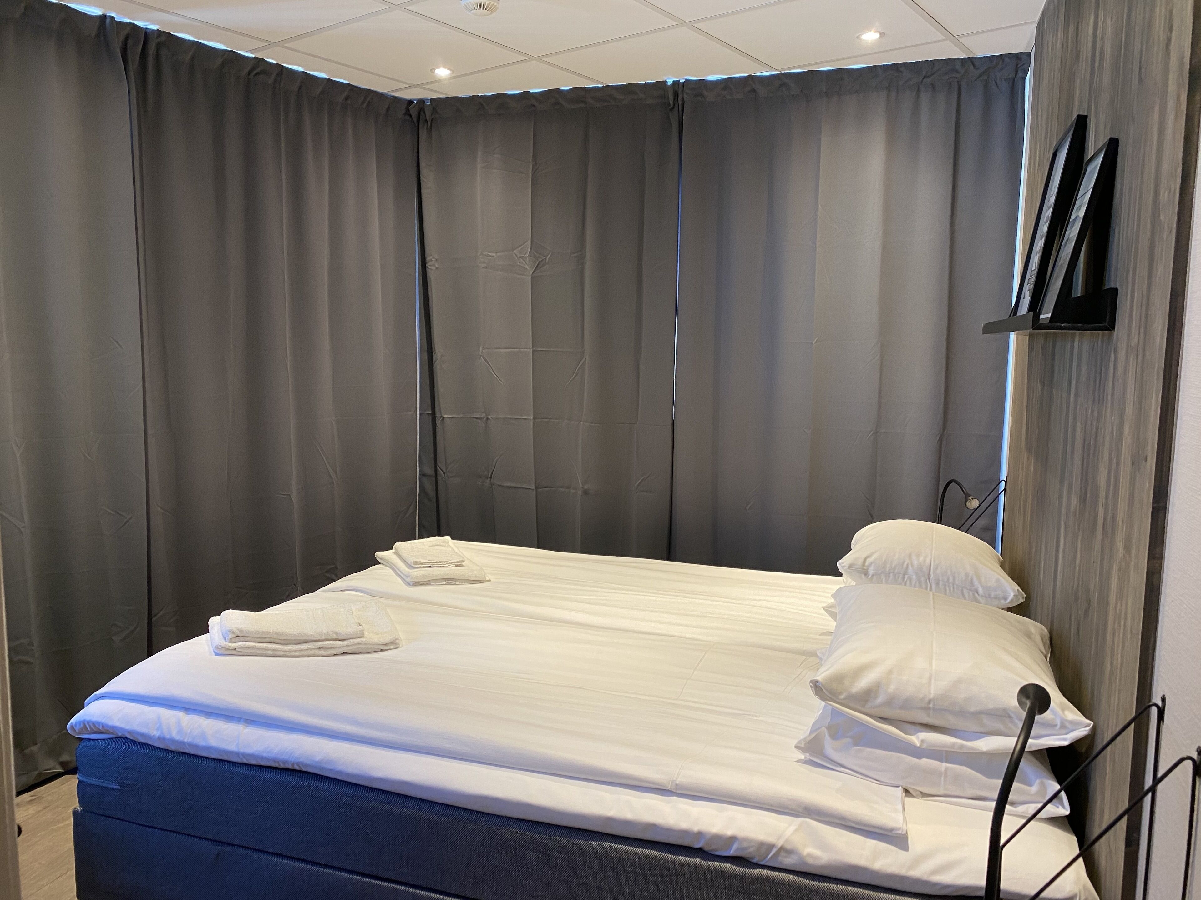 Cama bem arrumada com lençóis e almofadas brancos, cortinas cinzas e cabeceira de madeira.