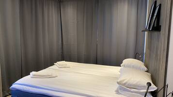 Cama bem arrumada com lençóis e almofadas brancos, cortinas cinzas e cabeceira de madeira.