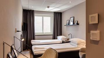 Une chambre d’hôtel avec deux lits simples, un bureau avec une chaise, une fenêtre avec des rideaux, une étagère avec des photos encadrées et un luminaire au plafond.