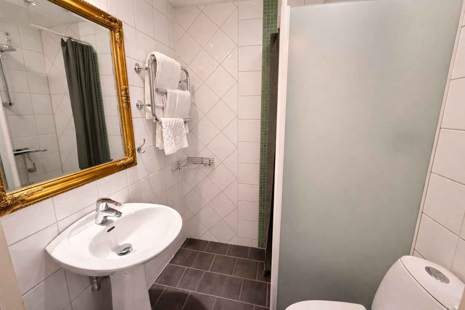 Un baño con lavamanos blanco, un espejo con marco dorado, toallero y cabina de ducha.