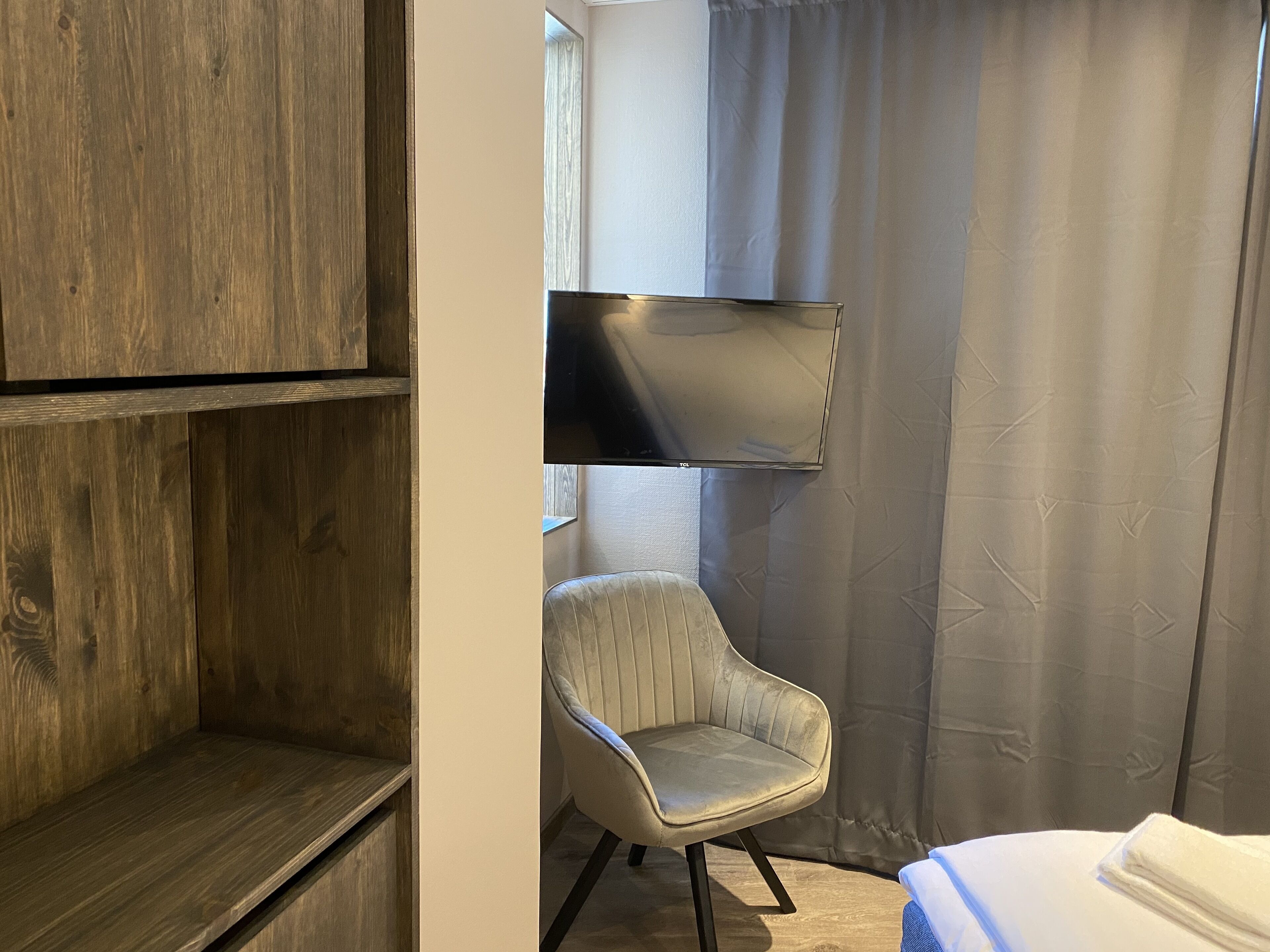 Ein Zimmer mit einem Holzschrank, einem grau gepolsterten Sessel, einem Bett mit weißer Bettdecke und einem grauen Vorhang.