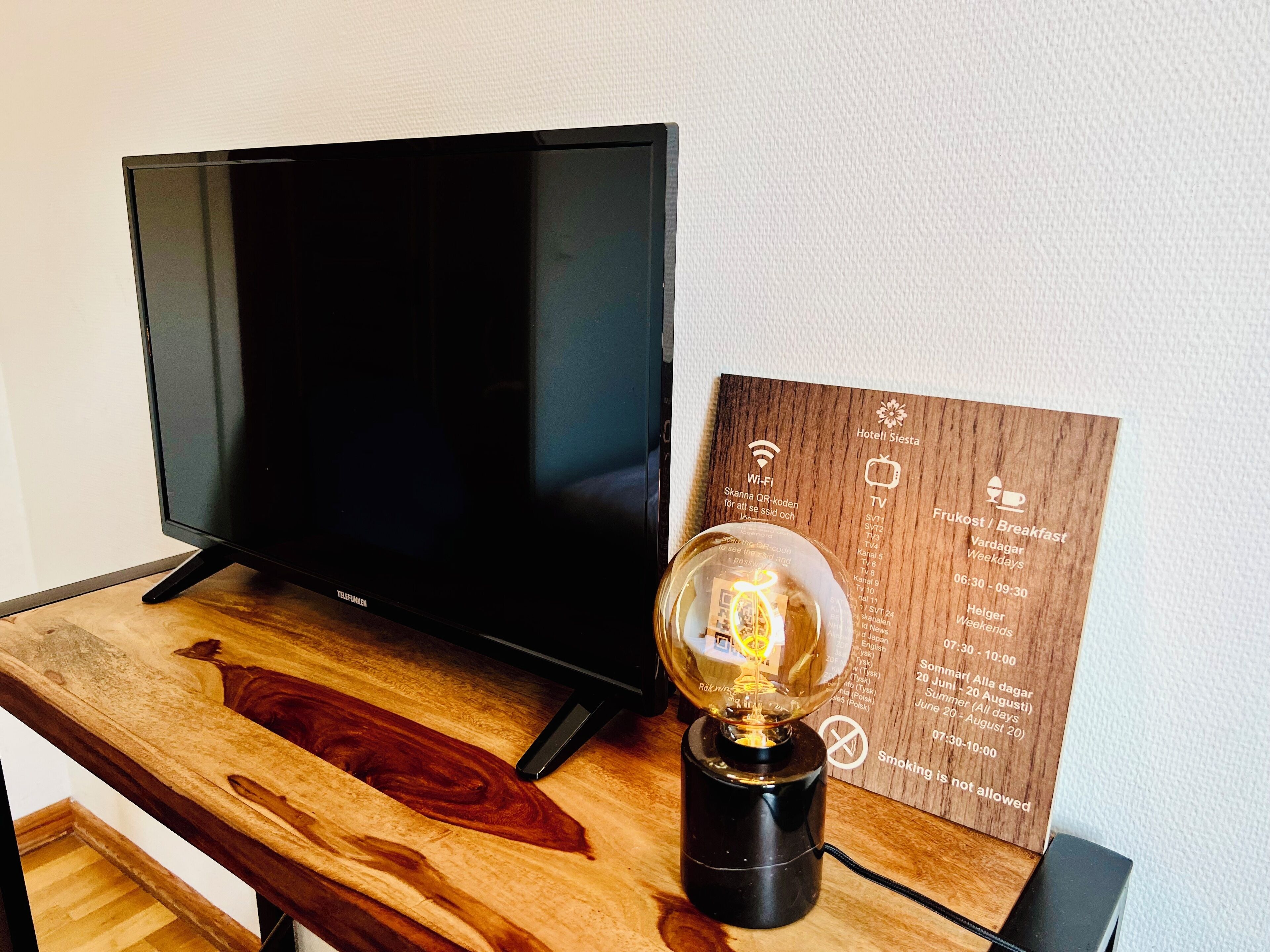Un televisor de pantalla plana sobre una mesa de madera, con una lámpara de vidrio y un letrero de madera que detalla los servicios del hotel y la disponibilidad de Wi-Fi.