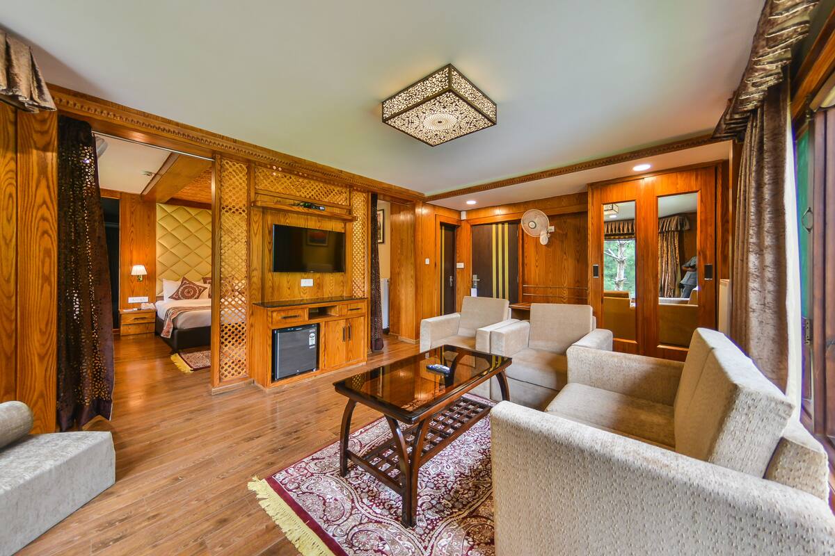 A spacious living room with a wooden floor, a glass coffee table, and white armchairs. There is a television and a small cabinet in the background.