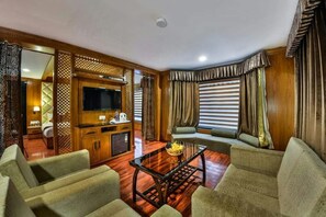 Quarto de hotel com decoração em madeira, TV de tela plana, sofá, mesa de centro e janela com cortinas.