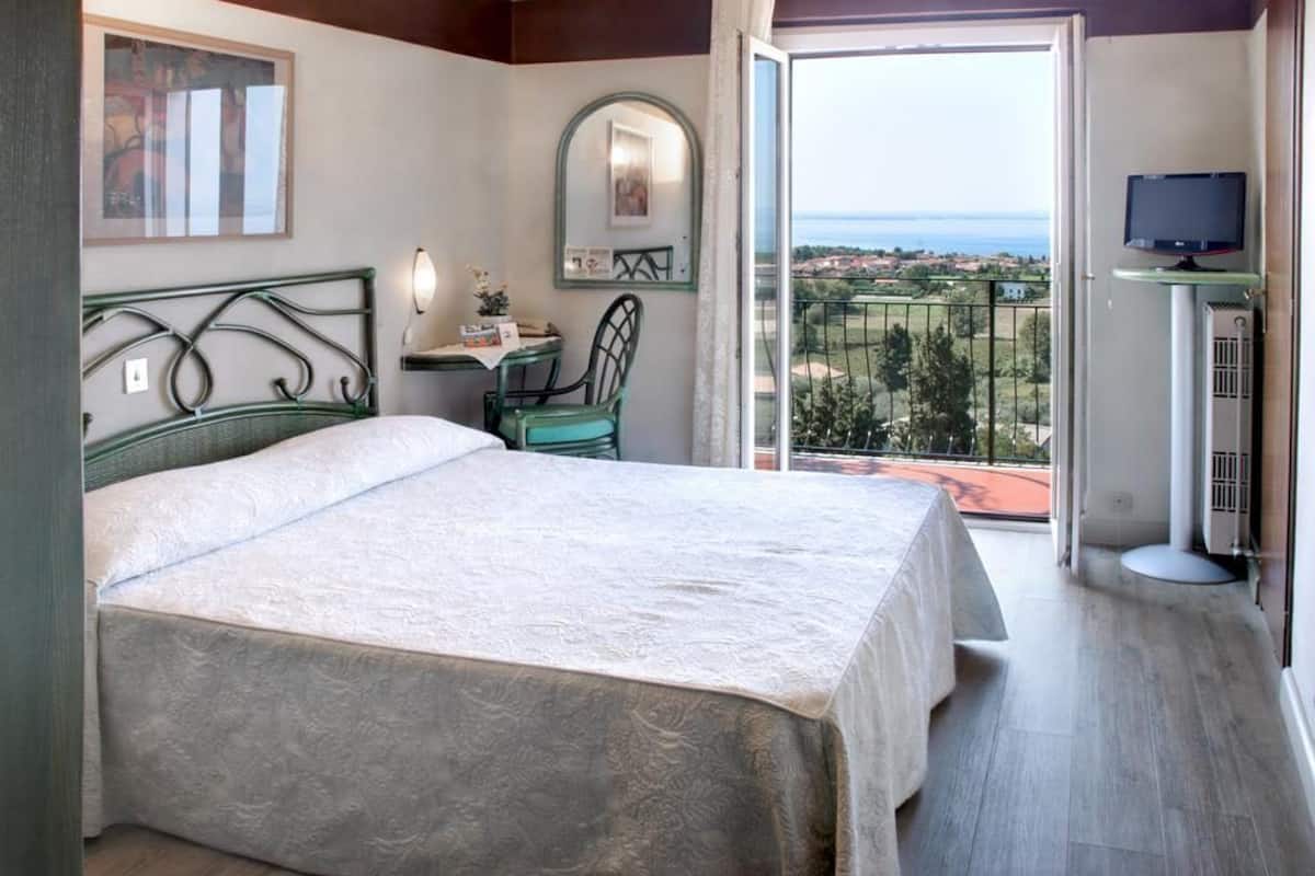 A bedroom with a bed, a desk, a chair, a TV, and a balcony with a view.