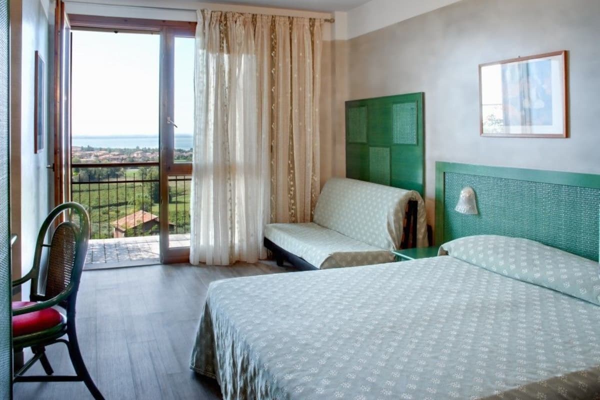A hotel room with two beds, a chair, a balcony with a view, and a green headboard.