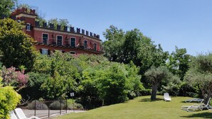 A red building with a balcony, surrounded by lush greenery and a well-maintained lawn. - Hotel Castello S. Antonio (Lazise)