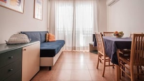 A small, neatly arranged room with a sofa, a dining table with chairs, a small cabinet, and a window with sheer curtains.