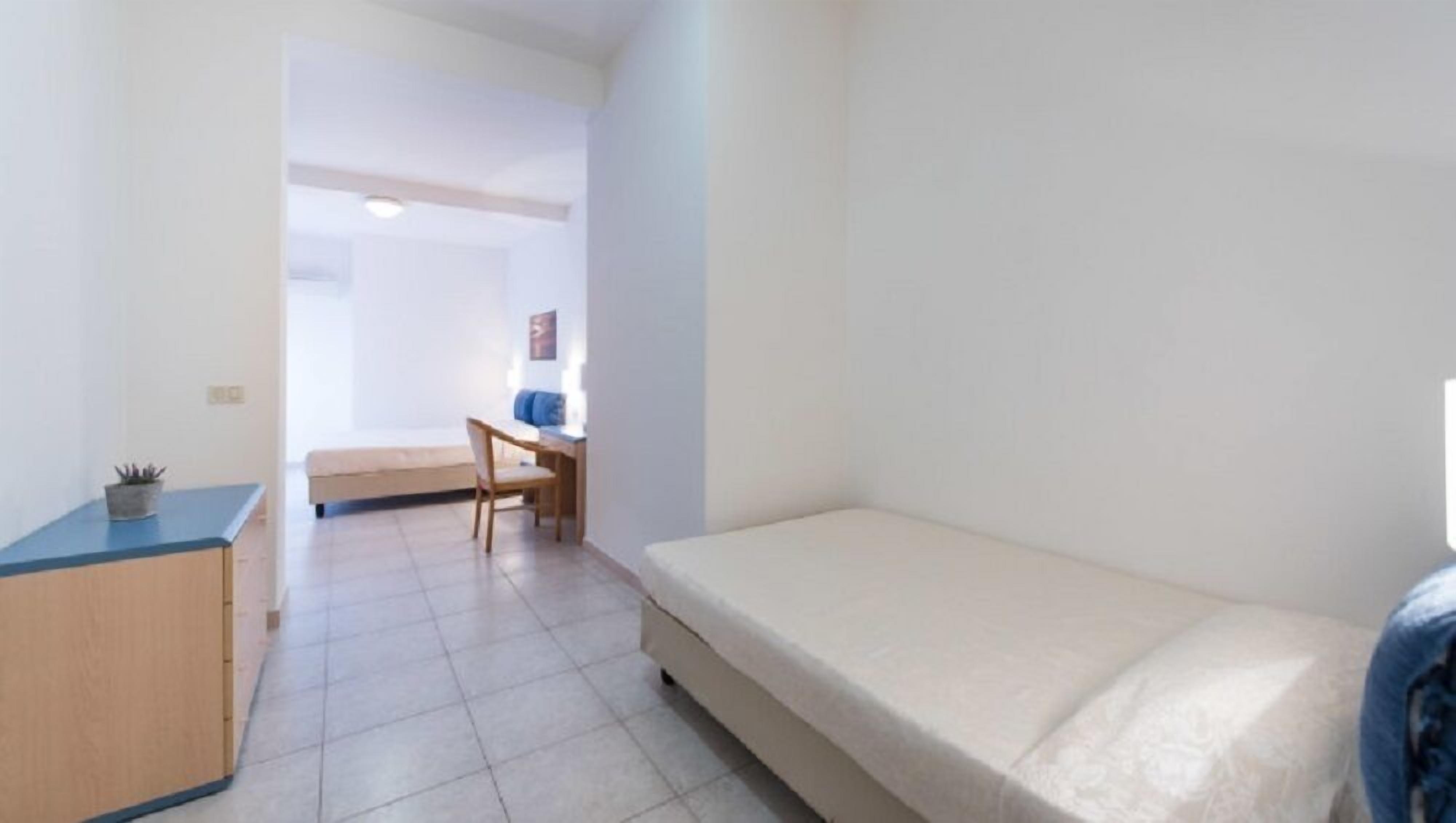 A small, sparsely furnished room with a bed, a desk, and a chair.