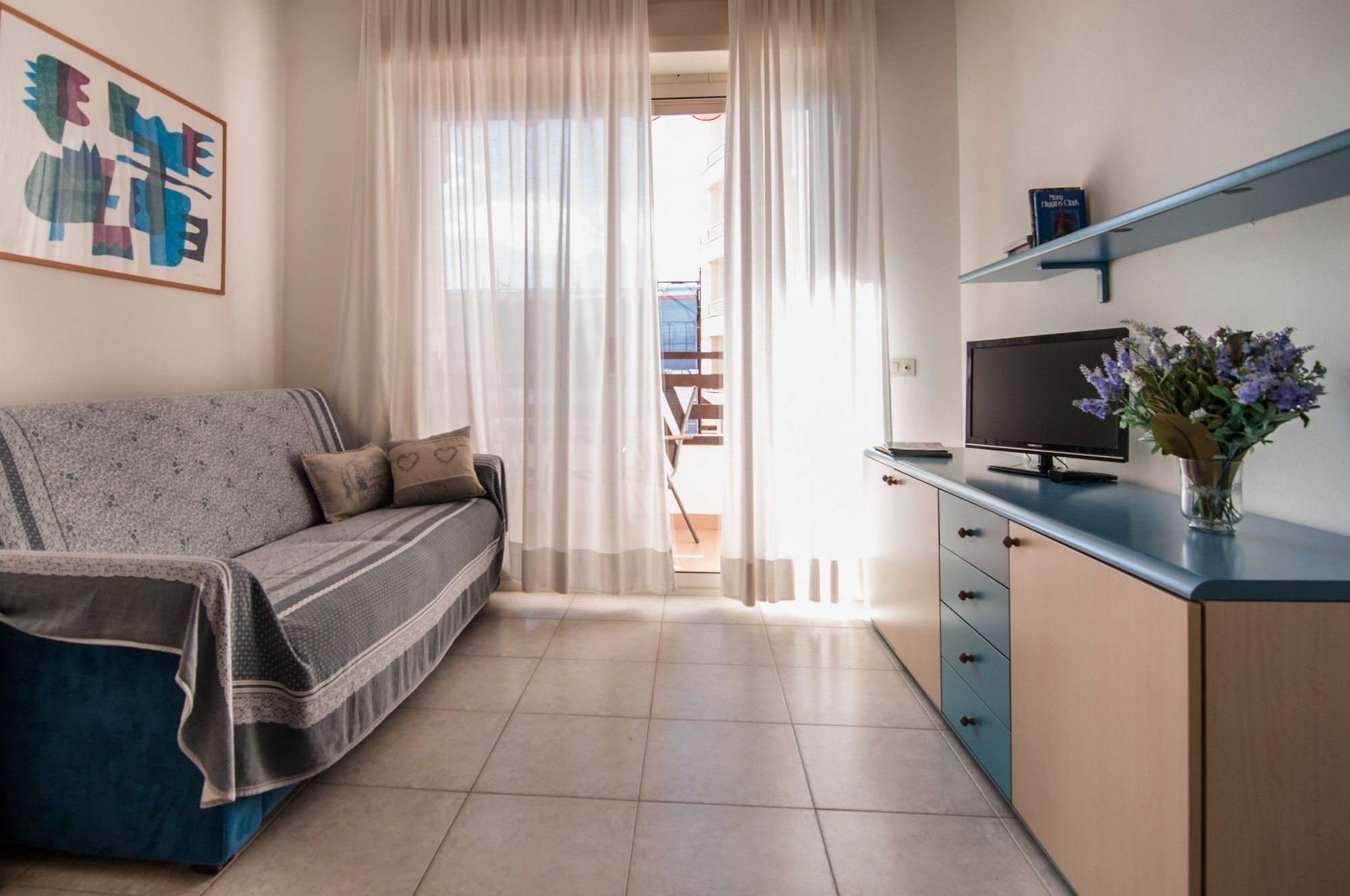 A modern living room with a sofa, a television on a cabinet, and a view through a window with sheer curtains.