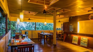 A restaurant with wooden flooring, tables, chairs, pendant lights, and a view of trees through large windows. - The Last Frontier Boutique Resort			 (Kota Kinabatangan)