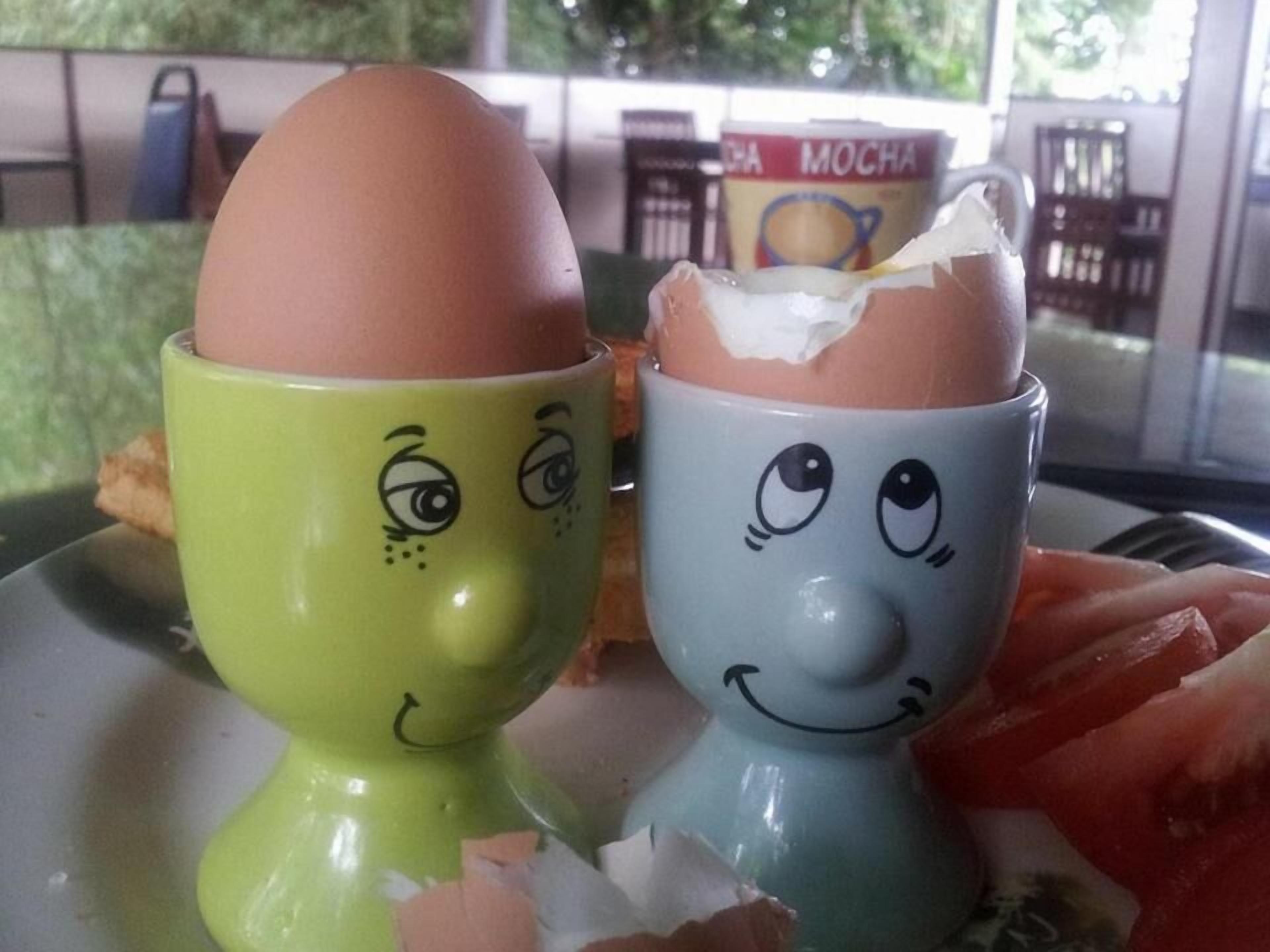 Two ceramic cups with faces, one green and one blue, each containing a cracked egg.