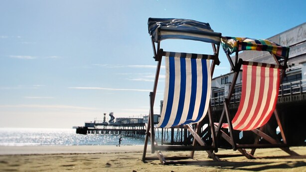 Two beach chairs with striped and solid colored canopies, a pier in the background, and a clear sky.