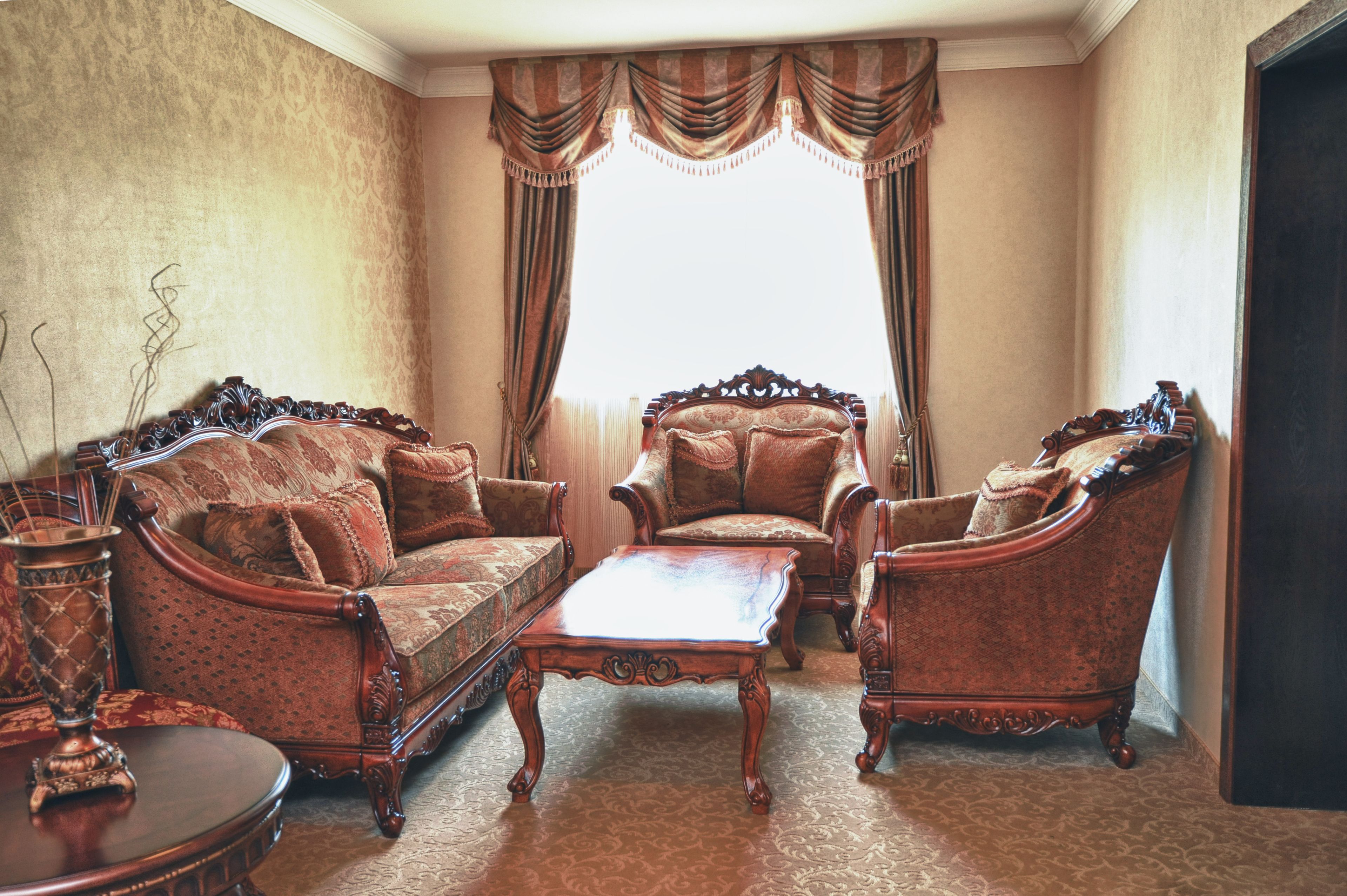 A traditional living room with ornate furniture, a patterned carpet, and a large window with curtains.