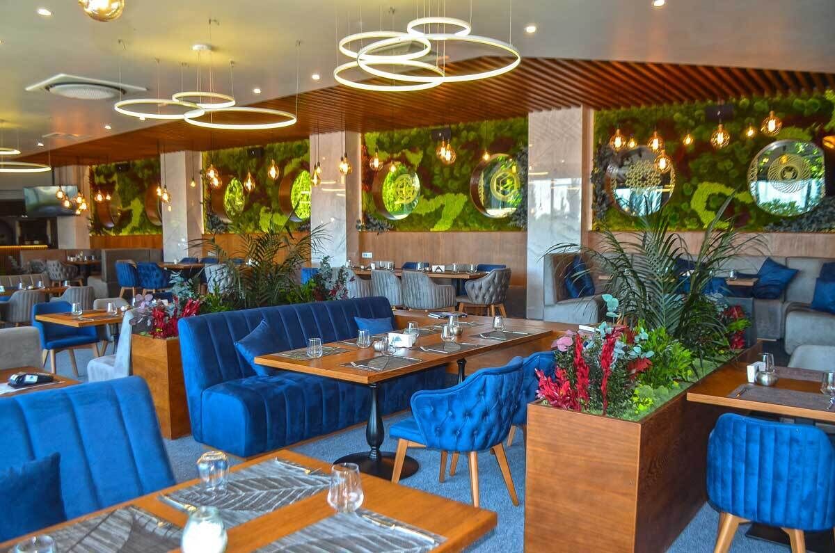 A modern restaurant with blue upholstered seating, wooden tables, and a green wall with circular mirrors.