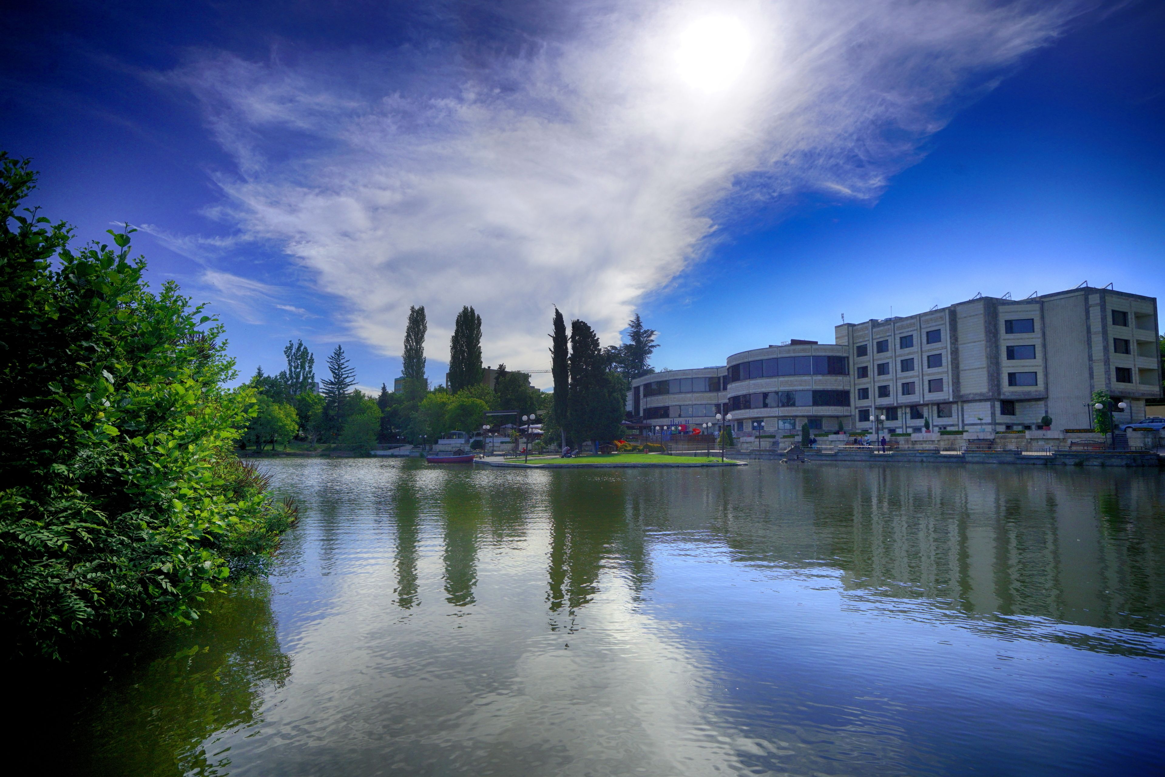 A calm lake with reflections, surrounded by greenery and buildings.