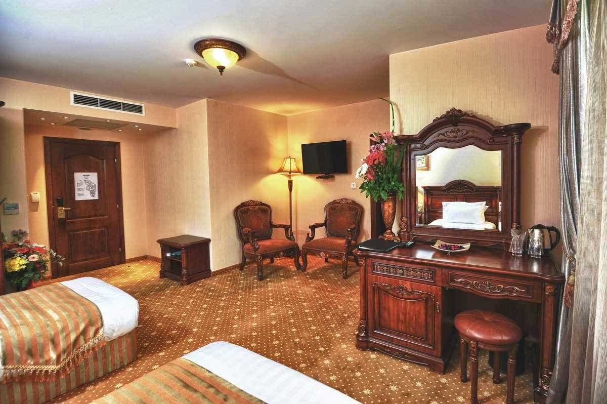 A hotel room with a bed, two chairs, a desk with a mirror, a lamp, and a television on the wall.