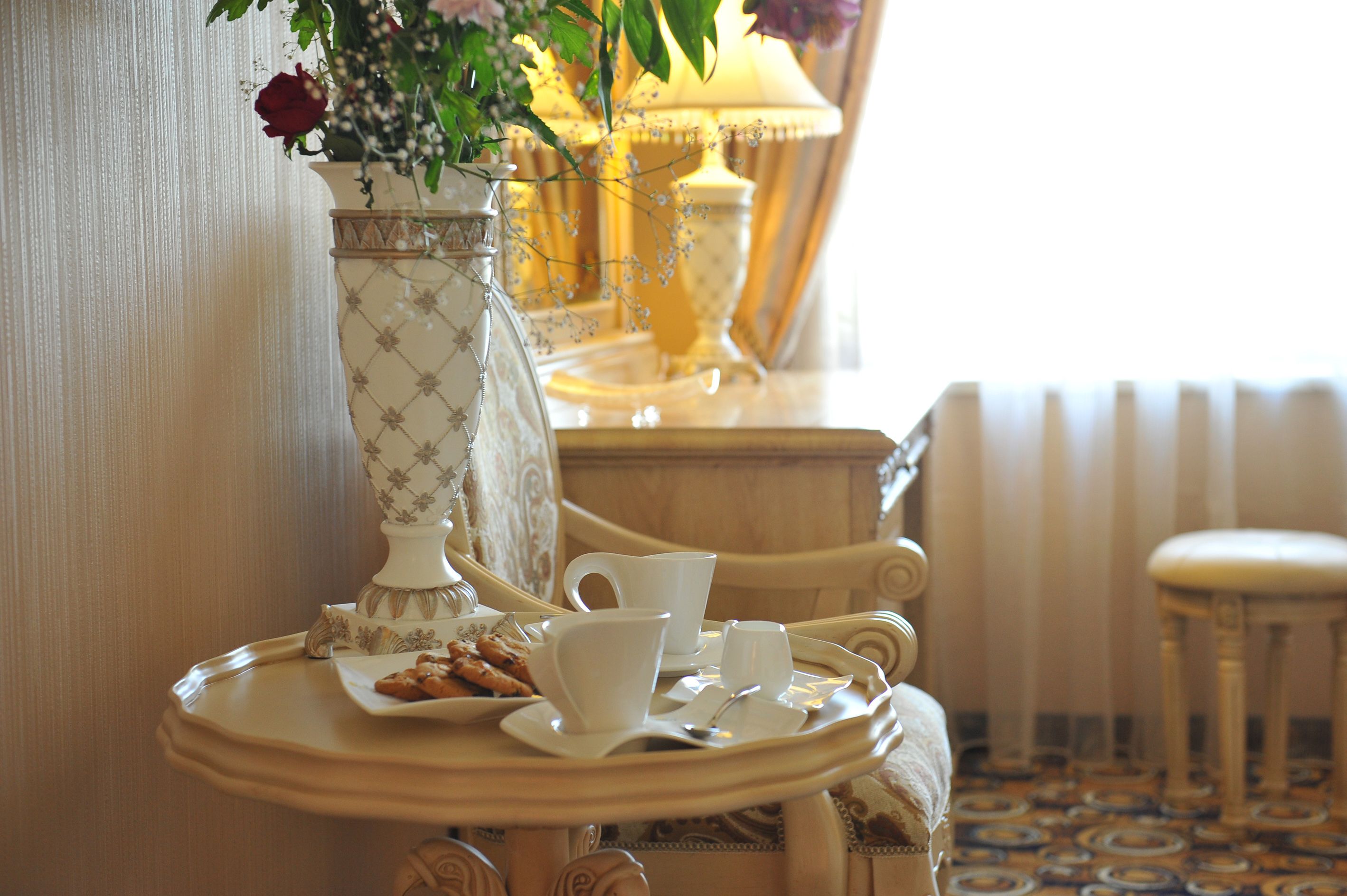 A dining table with a tray containing a teapot, cups, and pastries, set against a backdrop of a lamp and a vase with flowers.