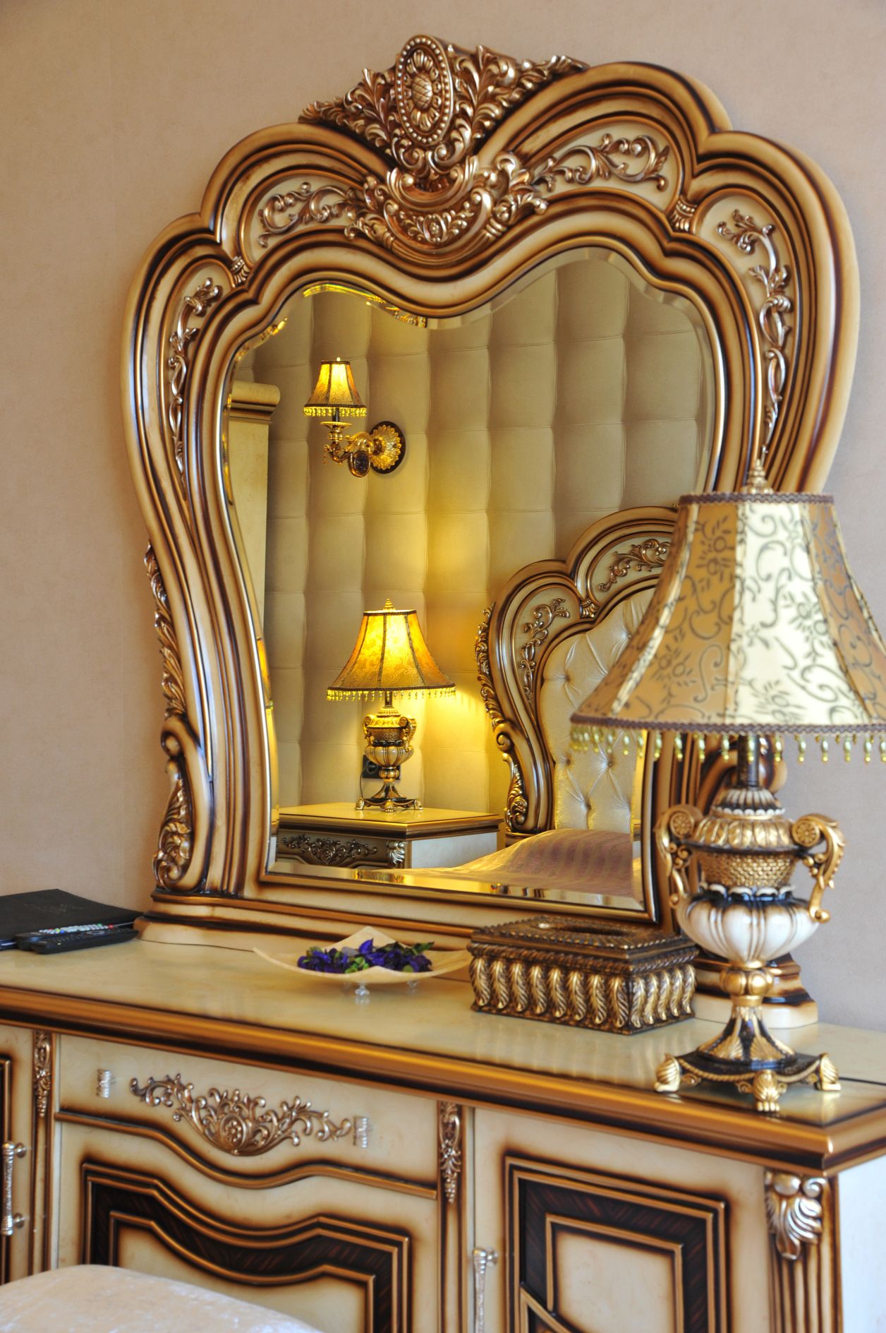 Een luxe wastafelmeubel met een sierlijke spiegel met gouden lijst, een bijpassende lamp en een decoratieve schaal op een beige gestoffeerde console.
