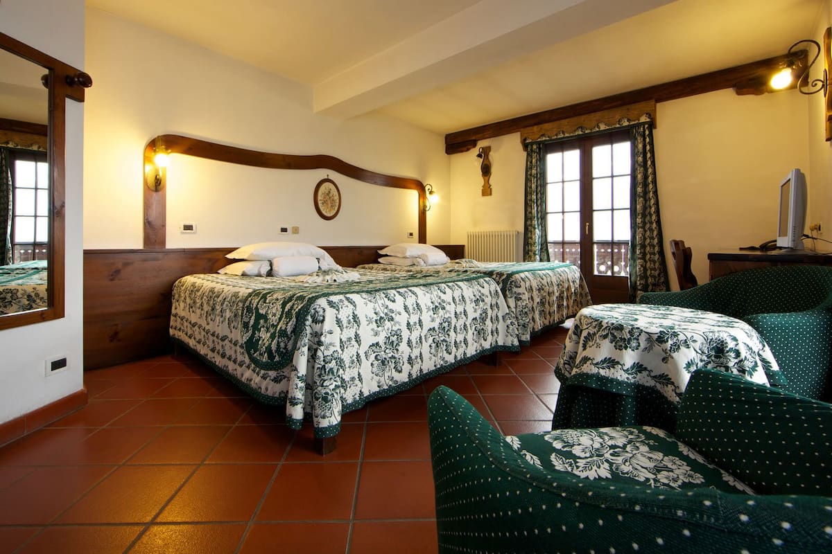 A hotel room with two beds, a wooden headboard, a large window with curtains, a small table with a television, and a chair with a floral cushion.