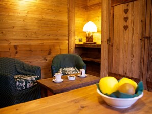 Ein gemütliches Holzhütten-Zimmer mit zwei Stühlen, einem Tisch mit einer Kaffeetasse und einer Obstschale, sowie einer Lampe auf einem Regal.