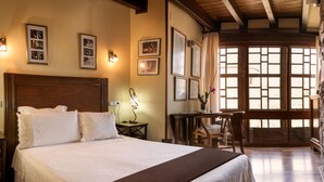 Hotelkamer met een groot bed, houten hoofdeinde, een bureau met stoel, en een raam met houten luiken.