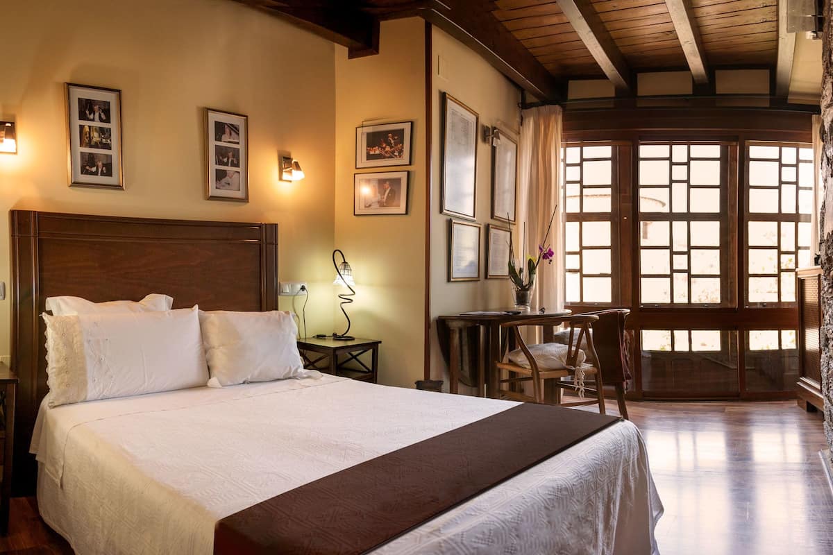 Hotelkamer met een groot bed, houten hoofdeinde, een bureau met stoel, en een raam met houten luiken.