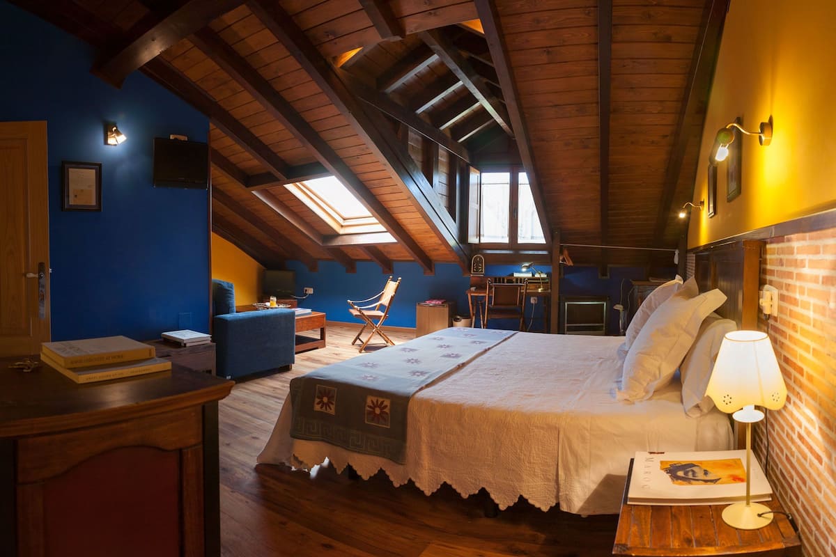 Een gezellige slaapkamer onder de dakpannen, met een houten plafond, een bed met een witte spreideken, een bureau met een computer en een blauwe fauteuil.