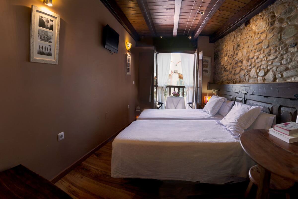 Een slaapkamer met twee bedden, een stenen muur, een houten plafond en een balkon met een tafel en stoelen.