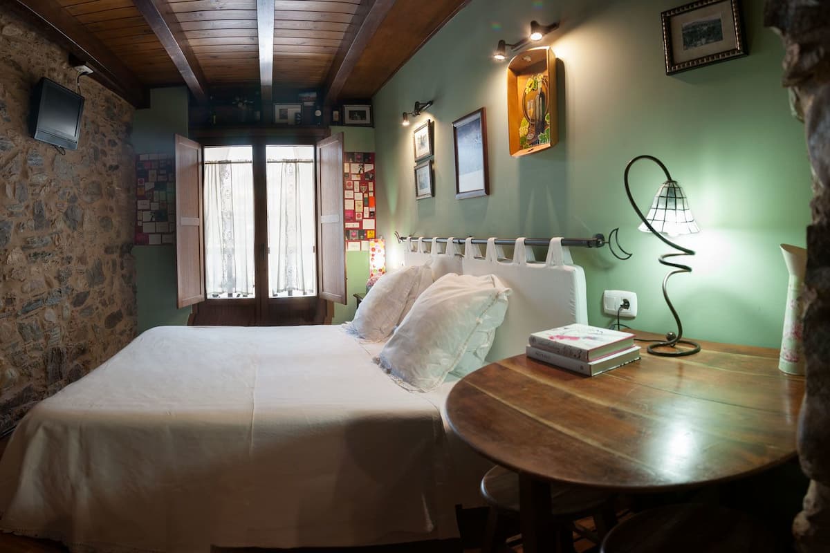 Een slaapkamer met een stenen muur, een bed met witte lakens, een ronde houten tafel met een boek en een raam met gordijnen.