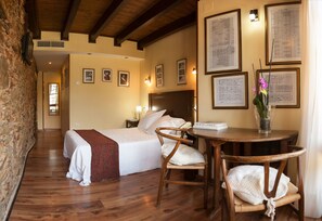 A hotel room with a wooden floor, a bed with white linens, a wooden chair, a small round table with a book and a vase of flowers, and framed pictures on the wall.