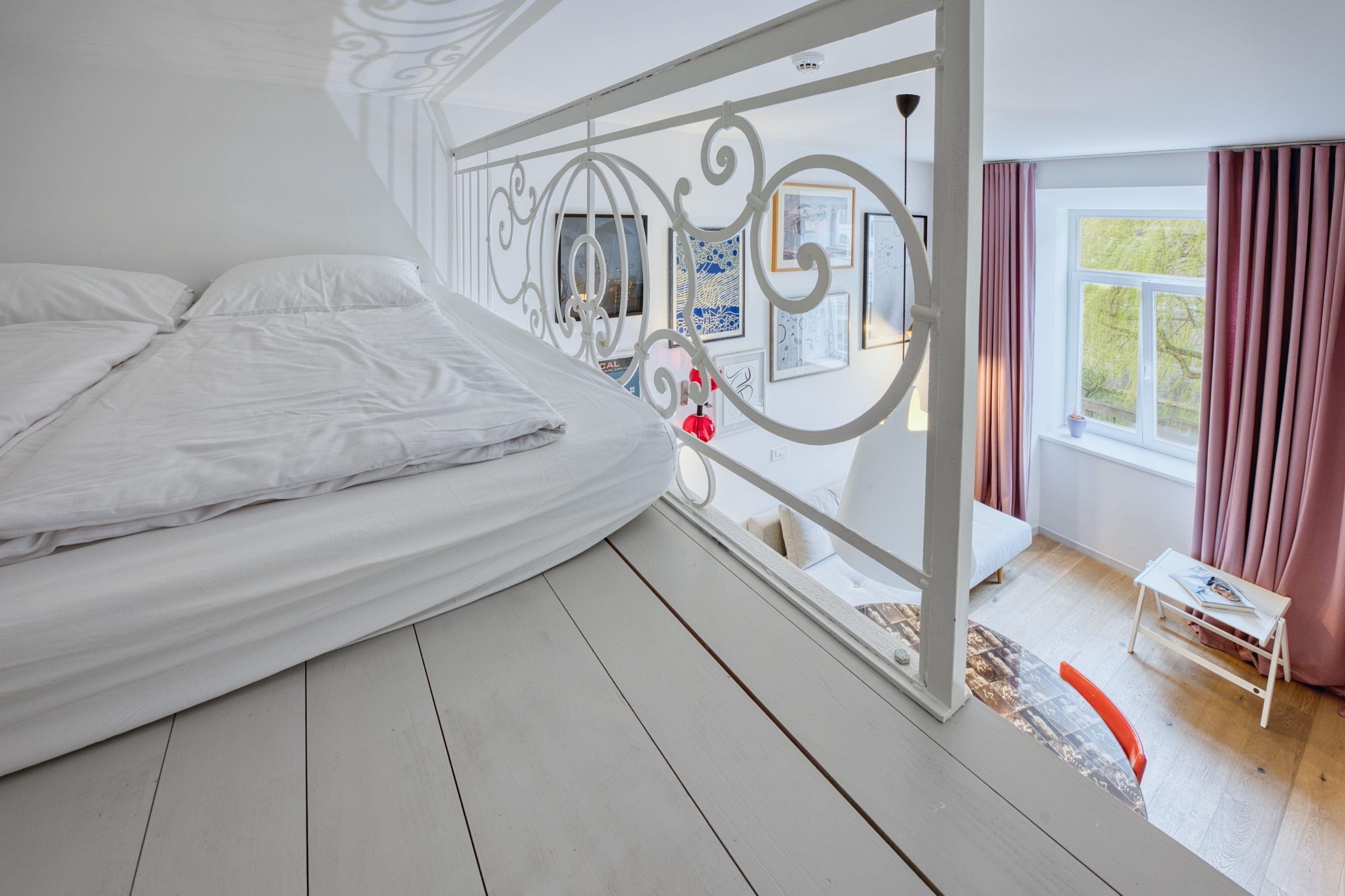 A bedroom with a bed, a window with curtains, a small table with a lamp, and a decorative metal railing.